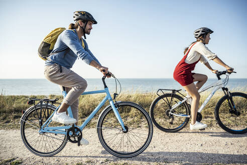 Couple wearing helmet riding bicycles against sea - UUF22224