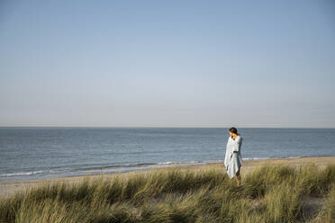 Junge Frau mit Kopftuch, die am Strand stehend die Aussicht betrachtet - UUF22225