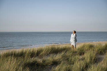 Young woman wrapped in shawl standing at beach - UUF22226