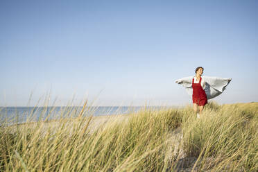 Junge Frau mit Kopftuch, stehend mit ausgestreckten Armen, läuft am Strand - UUF22229