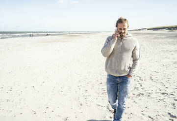 Young man talking on mobile phone while walking with hand in pocket at beach - UUF22233