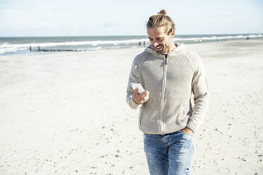 Mann mit Hand in der Tasche benutzt sein Smartphone beim Strandspaziergang - UUF22236