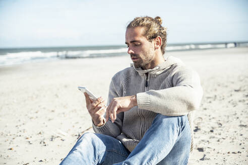 Mann, der sein Smartphone benutzt, während er an einem sonnigen Tag am Strand sitzt - UUF22238