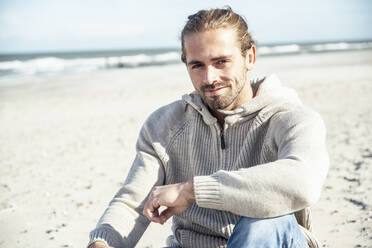 Smiling man sitting with hand on knee at beach - UUF22239