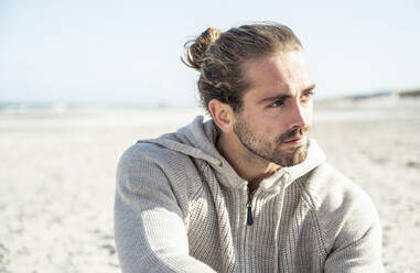 Junger Mann mit Kapuzenhemd, der am Strand sitzt und wegschaut, während die Sonne scheint - UUF22240
