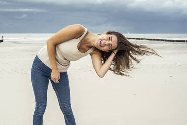 Cheerful woman with hand in hair standing at beach - UUF22248