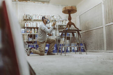 Handwerker mit Gesichtsschutzmaske beim Sprühen auf einem Tisch in der Werkstatt - OCAF00566
