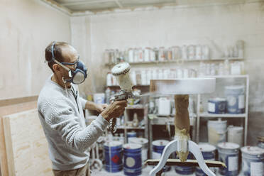 Handwerker mit Gesichtsschutzmaske, der einen Tisch mit einer Spritzpistole lackiert, während er in einer Werkstatt arbeitet - OCAF00574