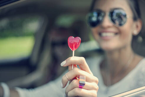 Happy young woman showing heart shape lollipop during road trip - AJOF00820