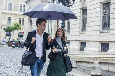 Geschäftsfrau mit männlichem Kollegen, die unter einem Regenschirm auf einer Straße in der Stadt spazieren gehen - AJOF00831