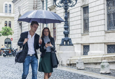 Businesswoman using smart phone while walking with male colleague on street in city - AJOF00832