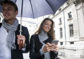 Lächelnde Geschäftsfrau, die unter einem Regenschirm mit einem männlichen Kollegen in der Stadt während der Regenzeit Textnachrichten auf einem Smartphone verschickt - AJOF00833