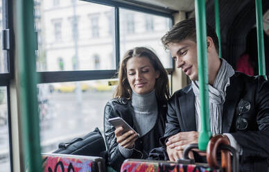 Lächelnde weibliche und männliche Kollegen, die ihr Smartphone beim Pendeln im Bus benutzen - AJOF00838