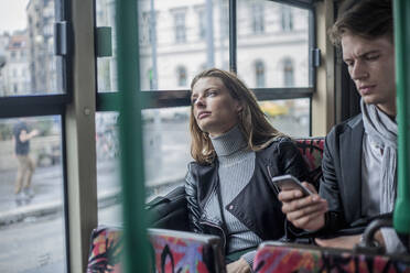 Nachdenkliche Frau, die mit einem männlichen Kollegen im Bus sitzt und ein Smartphone benutzt - AJOF00839