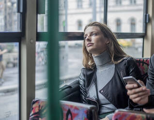 Nachdenkliche junge Geschäftsfrau, die im Bus neben einem männlichen Kollegen sitzt und sein Smartphone in der Hand hält - AJOF00840