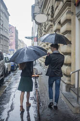 Männliche und weibliche Kollegen, die in der Regenzeit mit dem Fahrrad auf dem Bürgersteig spazieren gehen - AJOF00841