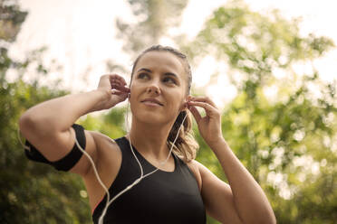 Sportswoman adjusting in-ear headphones while standing outdoors - AJOF00842