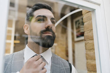 Young groom adjusting necktie while looking through window at home - EGAF01123