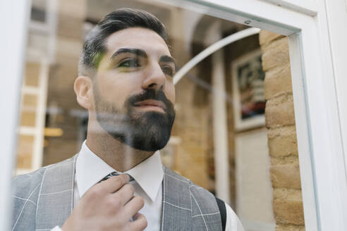Junger Bräutigam passt seine Krawatte an, während er durch das Fenster seines Hauses schaut - EGAF01123