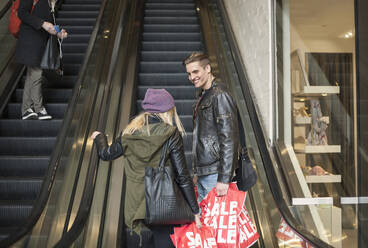 Junges Paar mit Einkaufstaschen auf einer Rolltreppe in einem Einkaufszentrum - AJOF00875