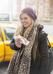Blonde Frau hält Hotdog, während sie auf der Straße in der Stadt steht - AJOF00878