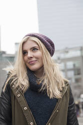 Smiling blond woman wearing knit hat looking away while standing in city - AJOF00881