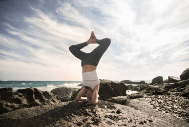 Woman practicing Sirsasana on rock formation at beach against sky - AJOF00891