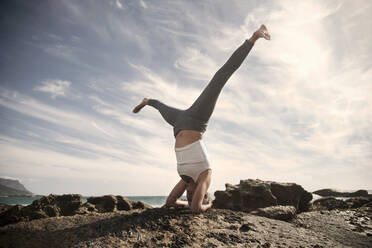Carefree woman practicing Sirsasana with legs apart on rock formation at beach against sky - AJOF00892