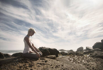 Frau meditiert bei der Ausübung von Yoga im Lotussitz auf einer Felsformation am Strand - AJOF00893