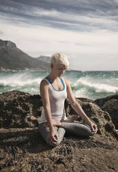 Blonde Frau meditiert beim Yoga im Lotussitz auf einer Felsformation am Strand - AJOF00894