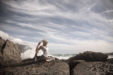 Flexible blond woman practicing yoga on rock formation at beach - AJOF00898