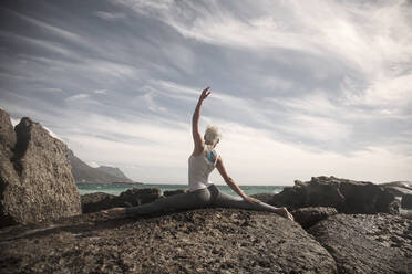 Woman with legs apart and hand raised practicing yoga on rock formation - AJOF00902
