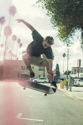 Carefree young man flipping skateboard in mid-air on street - AJOF00937