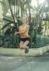 Happy young woman skipping with jump rope over sidewalk in morning - AJOF00949