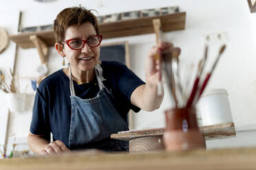 Smiling artist choosing paint brush in ceramic workshop - EGAF01175