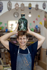 Lächelnde Künstlerin mit türkisfarbener Flasche auf dem Kopf im Keramikladen - EGAF01192