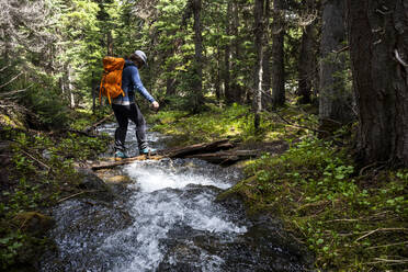 Side view of unrecognizable hiker with backpack crossing narrow fast stream flowing through green woods in mountainous region - CAVF91339