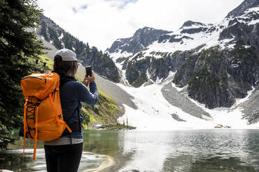 Eine Wanderin mit Rucksack und Smartphone steht in der Nähe eines Sees und fotografiert die malerische Landschaft der schneebedeckten felsigen Berge während einer Sommerreise in British Columbia - CAVF91340