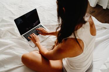 Von oben Rückenansicht einer nicht erkennbaren Frau in Schlafkleidung, die auf dem Bett sitzt und nach dem Aufwachen in einem hellen Schlafzimmer am Morgen in einem Laptop surft - ADSF18560