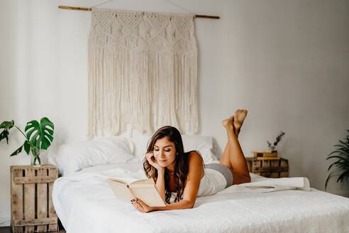 Positive junge Frau in Unterwäsche lehnt auf einem gemütlichen Bett und liest ein Buch, während sie den Wochenendmorgen in einem hellen Schlafzimmer mit Boho-Dekoration verbringt - ADSF18572