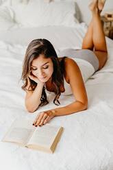 Positive junge Frau in Unterwäsche lehnt auf einem gemütlichen Bett und liest ein Buch, während sie den Wochenendmorgen in einem hellen Schlafzimmer mit Boho-Dekoration verbringt - ADSF18576