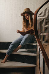 Pensive female wearing hat and jeans sitting on stairs in modern house and reading interesting book while relaxing at weekend at home - ADSF18581