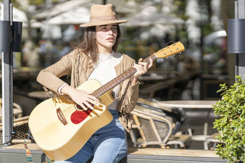 Junge Frau spielt Gitarre, während sie an einem sonnigen Tag auf einer Bank sitzt - GGGF00255