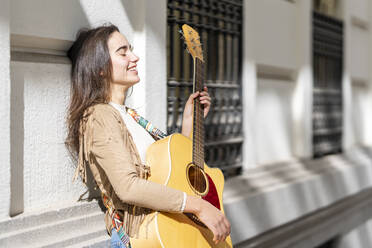 Smiling female musician playing guitar in city on sunny day - GGGF00268