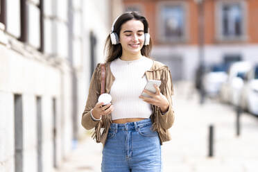 Smiling woman with headphones using smart phone on footpath - GGGF00271