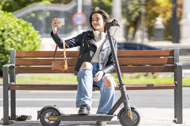 Smiling woman taking selfie through smart phone while sitting on bench by electric push scooter - GGGF00279