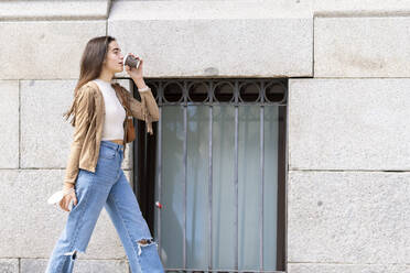 Young woman drinking coffee while walking by wall - GGGF00294