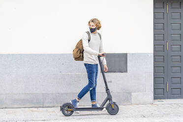 Young man wearing protective face mask standing with electric push scooter on footpath - GGGF00297