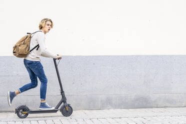 Mann mit Rucksack, der mit einem elektrischen Roller auf dem Gehweg gegen eine Mauer fährt - GGGF00300