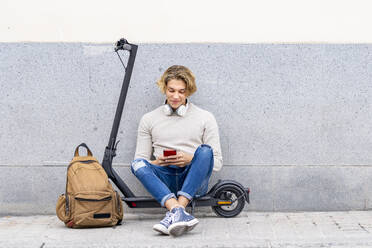 Smiling man with backpack using smart phone while sitting on electric push scooter - GGGF00302